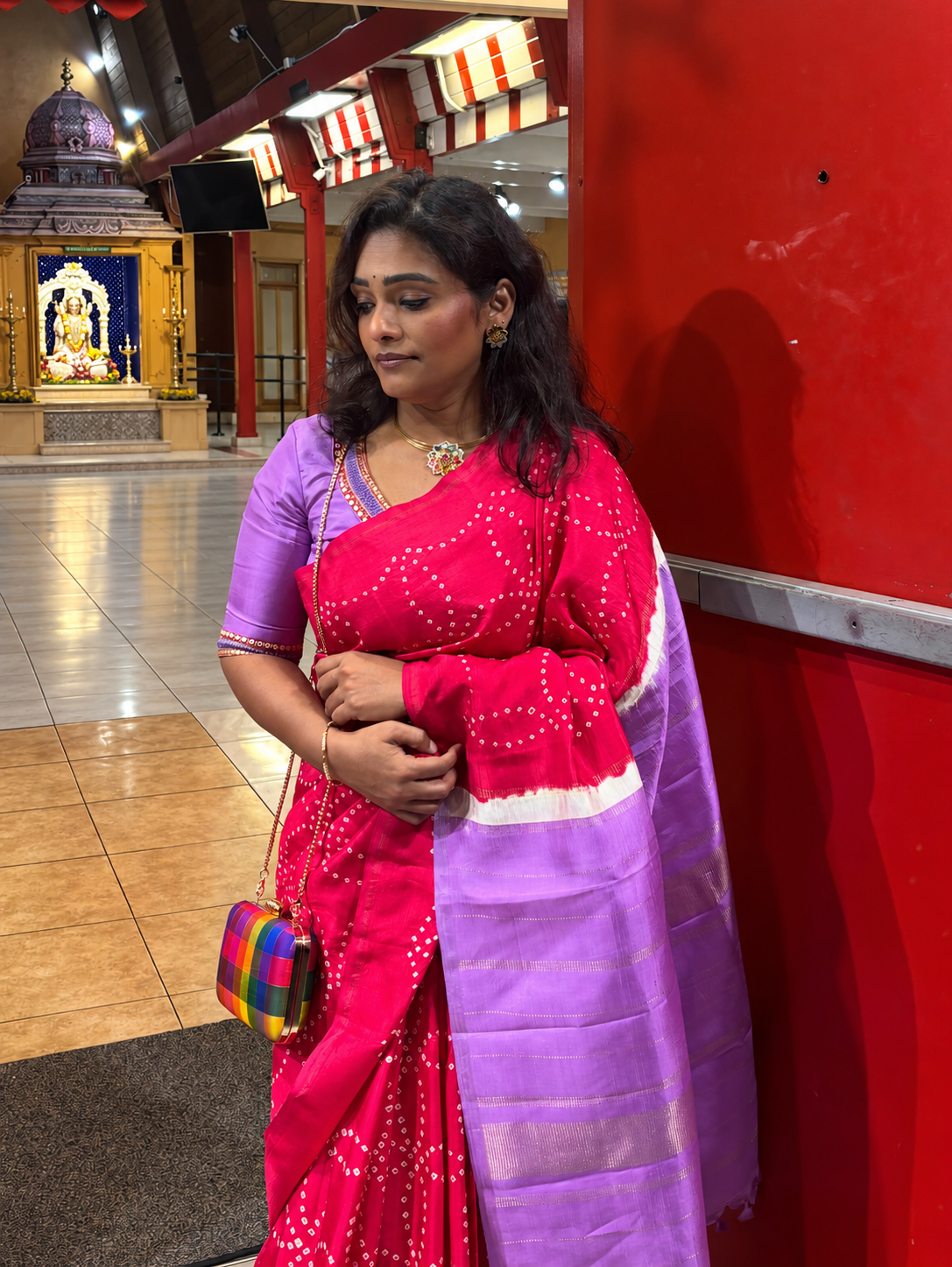 Subha beside a red temple door, wearing a pink saree and lavender blouse with a checked clutch